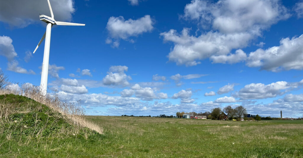Ett gräsfält med en gård längst bort vid horisonten. Blå himmel med små vita moln. En kulle med ett vindkraftverk i kanten av fältet.